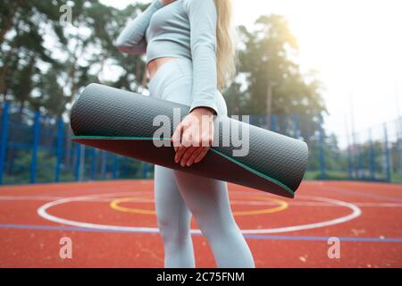 Concept de forme physique, de sport et de mode de vie sain. Image courte de jeune fille en vêtements de sport gris, tenant un tapis d'exercice et marchant à l'extérieur du sport Banque D'Images