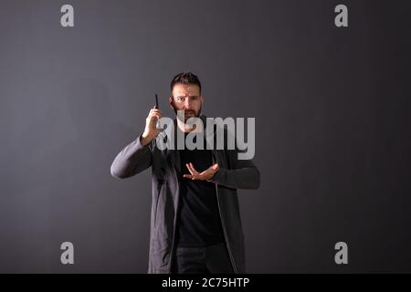 Un beau jeune homme dans un sweat gris en conversation sur son téléphone devant un fond gris foncé dans un studio. Banque D'Images