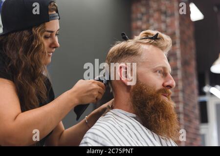 Le masque de coiffeur féminin coupe les poils d'un homme avec une tondeuse à cheveux. Coiffure pendant la prise de distance sociale Banque D'Images