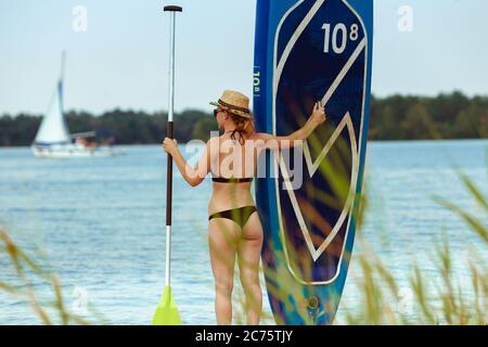 Jeune femme attrayante debout à côté du paddle board, SUP. Vie active, sport, loisirs concept. Femme caucasienne à bord en été soir. Vacances, station, plaisir. Banque D'Images