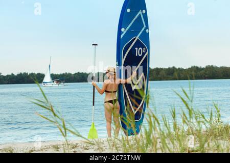 Jeune femme attrayante debout à côté du paddle board, SUP. Vie active, sport, loisirs concept. Femme caucasienne à bord en été soir. Vacances, station, plaisir. Banque D'Images