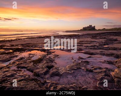 Un lever du soleil se reflète dans de beaux bassins de marée sur la plage de Bamburgh avec le célèbre château surplombant la scène sur l'horizon. Banque D'Images