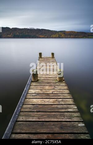 Une jetée en bois provient de la rive du Windermere dans le Lake District sous un ciel couvert journée d'automne, avec des collines boisées à l'arrière-plan. Banque D'Images