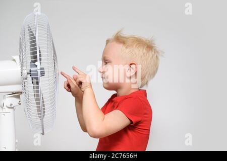 Un petit garçon blond dans un t-shirt rouge touche le ventilateur avec son doigt. Concept été Banque D'Images