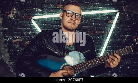 Homme tatoué jouant de la guitare près d'un mur éclairé. Un homme élégant avec tatouage oiseau sur la poitrine en regardant loin et jouant de la guitare tout en se tenant contre le chabby Banque D'Images