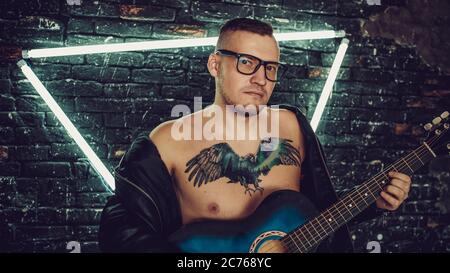 Homme tatoué jouant de la guitare près d'un mur éclairé. Un homme élégant avec tatouage oiseau sur la poitrine en regardant loin et jouant de la guitare tout en se tenant contre le chabby Banque D'Images