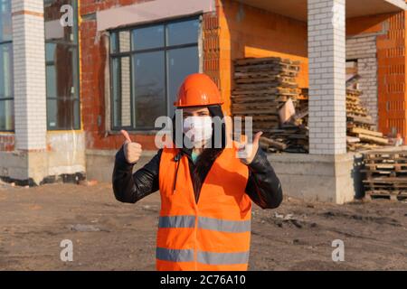Femme travailleur de la construction dans des salopettes et dans un masque médical montrant les pouces sur le fond de la maison en construction. Jeune femme en casque dur et Banque D'Images