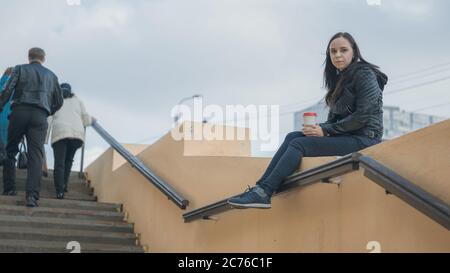 Une jeune femme belle en vêtements décontractés est assise sur une clôture en béton, regarde loin et tient une tasse de café. Banque D'Images