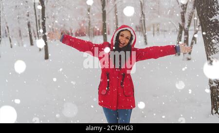 Portrait de la jeune femme en veste rouge et capuche en hiver. Belle dame se fait des vagues les mains et se réjouit de la chute de neige. Des enveloppements de neige moelleux Banque D'Images