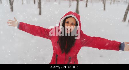 Portrait de la jeune femme en veste rouge et capuche en hiver. Belle dame se fait des vagues les mains et se réjouit de la chute de neige. Des enveloppements de neige moelleux Banque D'Images