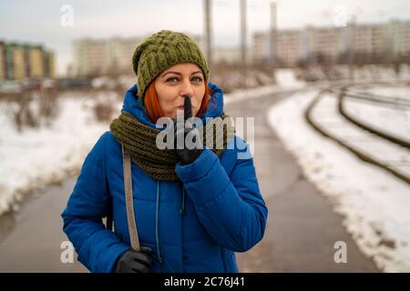 Belle jeune femme aux cheveux rouges dans des vêtements décontractés sourit et fait un geste de silence avec l'index sur ses lèvres. Le concept de silence. Banque D'Images