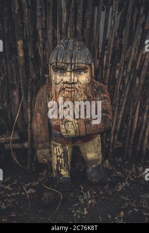 Gnome en bois près d'une clôture en bois au sol. Ancienne figure fissurée sculptée dans le bois. Gnome de jardin dans la campagne. Banque D'Images