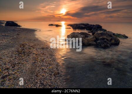 Tir en longue exposition pour capturer les vagues en mouvement sur une plage près de quelques rochers au premier plan contre le soleil levant avec des coquillages sur la côte tir en R. Banque D'Images