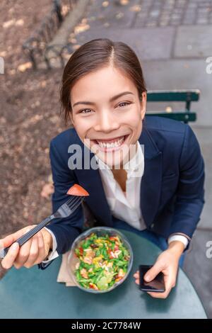 Pause déjeuner saine alimentation asiatique femme d'affaires prête à manger saladier dans City Park vie urbaine style de vie à l'aide du téléphone. Joyeux sourire multiracial Banque D'Images