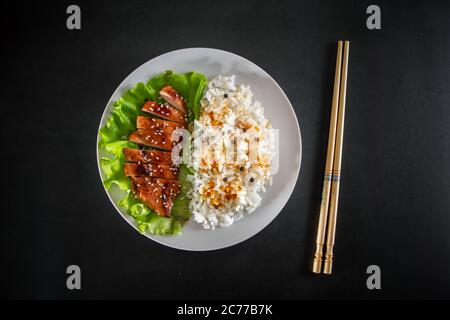 Poulet teriyaki avec riz blanc sur une assiette. Nourriture savoureuse et bâtonnets chinois sur fond sombre Banque D'Images