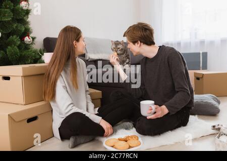 Portrait de jeune beau couple assis sur le sol à la maison avec des biscuits et regardant le chat près Banque D'Images