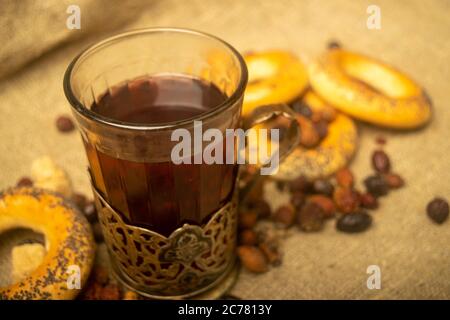 Un verre à facettes de thé dans un porte-gobelet vintage, des fruits de rosehip séchés et de petits bagels sur un fond de tissu de homespun avec une texture rugueuse. Gros plan Banque D'Images