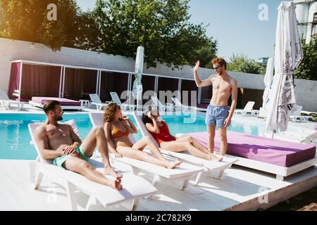 Trois jeunes hommes s'inclinent sur des chaises longues et un homme aux cheveux rouges se tenant à proximité Banque D'Images