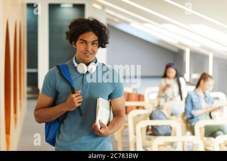 Portrait d'un beau garçon d'étudiant souriant avec des cheveux désordonnés debout avec cartable et ordinateur portable dans la salle universitaire moderne Banque D'Images