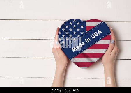 Inscription faite aux Etats-Unis, le drapeau des Etats-Unis. Mains de femmes tenant une boîte en forme de coeur. Table en bois blanc sur fond. Placer pour le texte Banque D'Images