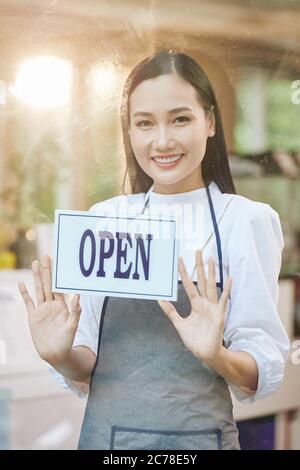 Portrait de la jolie jeune serveuse vietnamienne qui colle l'affiche sur la porte du café lors de l'ouverture de la porte après la période de verrouillage Banque D'Images