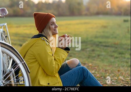 Belle femme souriante assise sur l'herbe dans le parc, tenant une tasse de café. Voyage, concept d'automne Banque D'Images