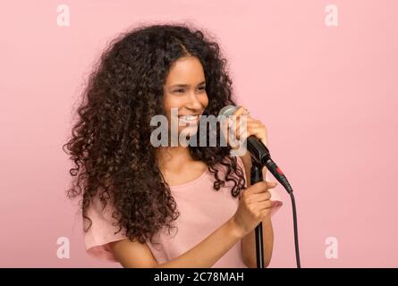 Vivacous jeune fille noire avec un microphone souriant heureux comme elle se tourne pour regarder sur le côté contre un fond rose studio Banque D'Images