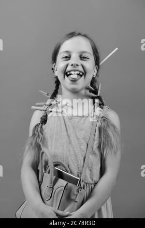 Enfant en robe rose avec des tresses et des crayons de couleur. Une fille tient le sac d'école dans les mains et le cookie en forme de coeur sur la langue. Fille d'école avec visage heureux isolée sur fond de saumon. Concept de retour à l'école Banque D'Images