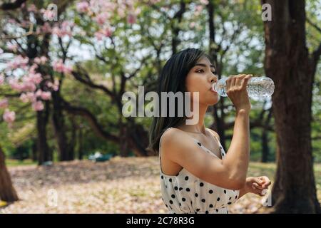 Jeune femme brune qui boit de l'eau dans le parc. Belle fille asiatique soif et prendre un verre d'eau à l'extérieur. Banque D'Images