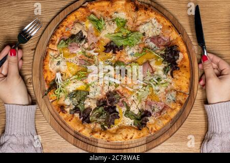 Grande pizza délicieuse et mains de femmes avec une fourchette et un couteau sur un fond en bois. Cuisine nationale italienne, restaurant Banque D'Images