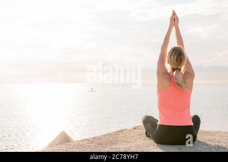 vue arrière d'une femme blonde méconnaissable pratiquant le yoga au lever du soleil, assise en face de la mer, concept de santé mentale et de détente, copie de spac Banque D'Images
