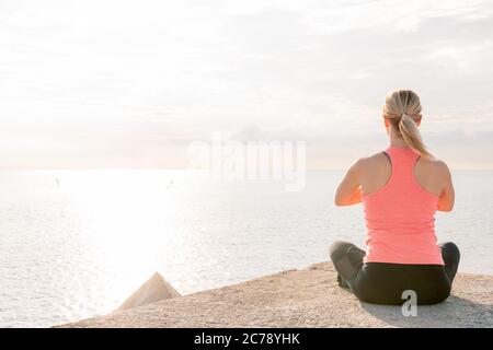 vue arrière d'une femme blonde méconnaissable faisant du yoga à l'aube, assise en face de la mer, concept de soins de santé mentale et de relaxation, espace de copie pour Banque D'Images