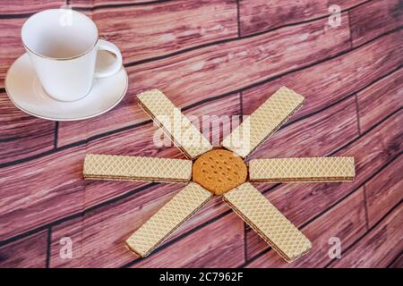 Composition avec un soleil fait de biscuits et de gaufrettes et une tasse d'espresso blanc sur une nappe avec des planches en bois Banque D'Images