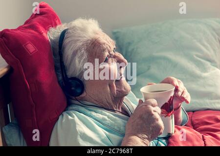 Care home âgée 95 ans dame heureuse de se reposer au lit portant des écouteurs apprécie une tasse de thé dans sa maison ensoleillée confortable de soins Banque D'Images