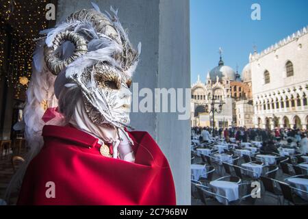 VENISE, ITALIE - 28 FÉVRIER 2019 : un élégant masque blanc avec un manteau rouge près d'une série de tables de bar pendant le carnaval de Venise Banque D'Images