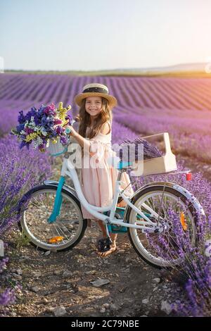 jolie petite fille avec fleurs et chapeau de paille ayant l'amusement dans le champ de lavande. Préadolescence fille avec vélo. Banque D'Images