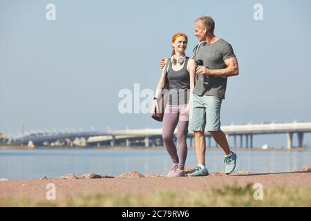 Homme mûr souriant avec une bouteille d'eau embrassant sa femme tout en marchant avec elle sur le bord de la rivière après l'entraînement Banque D'Images