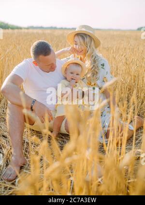 Une jeune famille heureuse avec bébé s'assoit et repose parmi le champ de blé jaune. Banque D'Images