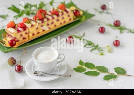 Tasse de café au lait, gâteau à la fraise, baies de cerise mûres et fleurs fraîches sur la table lumineuse. Petit déjeuner romantique d'été Banque D'Images