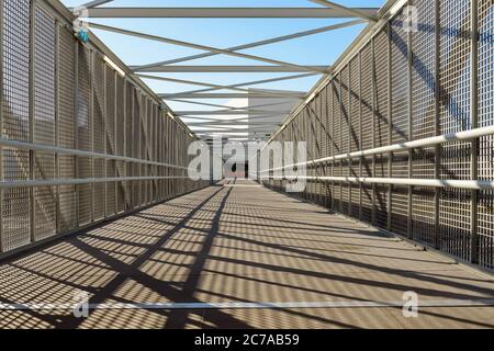 Intérieur d'un pont piétonnier moderne au-dessus d'une autoroute par temps ensoleillé. Diminution de la perspective et des ombres dues à la clôture sur le sol. Banque D'Images