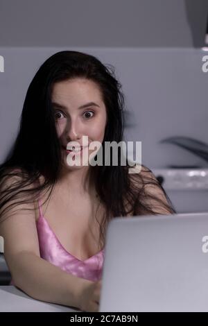 magnifique brune fille choquée en pyjama rose avec une tasse de thé-café bleu, travaille sur un ordinateur portable - travail à distance ou d'étude - dans une cuisine grise. Photo de haute qualité Banque D'Images