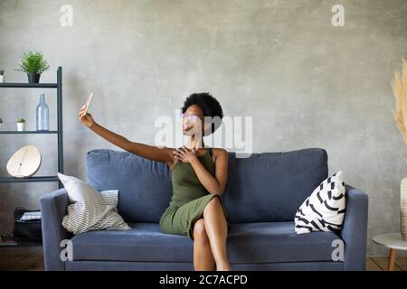 Bonne jeune femme afro-américaine s'assoit sur un canapé confortable et prend des selfies. Je vous souhaite un nouvel appartement. Banque D'Images