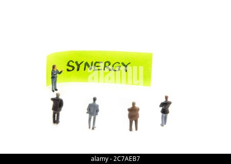 Réunion stratégique avec figurines miniatures posées comme des hommes d'affaires se tenant autour de post-it note avec message Synergy sur fond blanc, minimaliste a Banque D'Images