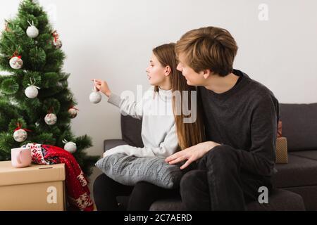 Portrait de beau jeune couple assis sur un canapé à la maison et décorant l'arbre de Noël ensemble Banque D'Images