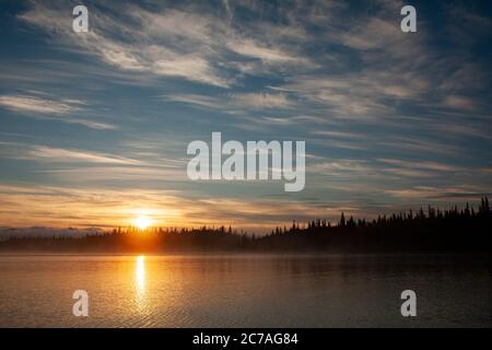 Coucher de soleil doré sur un lac d'Alaska, avec des silhouettes de montagne et des nuages doux reflétant la beauté sereine de la nature sauvage au crépuscule. Banque D'Images