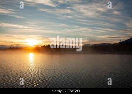 Coucher de soleil doré sur un lac d'Alaska, avec des silhouettes de montagne et des nuages doux reflétant la beauté sereine de la nature sauvage au crépuscule. Banque D'Images