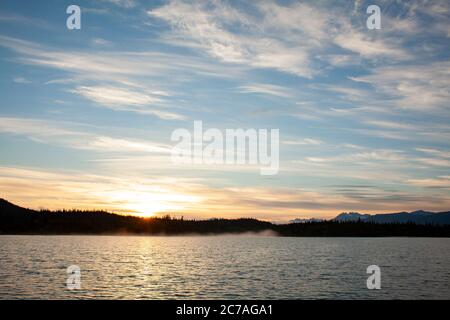 Coucher de soleil doré sur un lac d'Alaska, avec des silhouettes de montagne et des nuages doux reflétant la beauté sereine de la nature sauvage au crépuscule. Banque D'Images