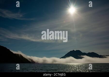 Le brouillard roule sur les eaux du lac Iliamna, en Alaska, avec le soleil projetant la lumière sur la silhouette accidentée de la montagne et le paysage sauvage serein Banque D'Images