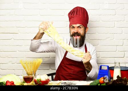 Concept de processus de cuisson. Cuisinez avec un visage excités en uniforme bordeaux, assis près d'une table avec des légumes et des ustensiles de cuisine. Chef fait de la pâte. Homme avec barbe étire la pâte à pétrir sur fond de brique blanche Banque D'Images