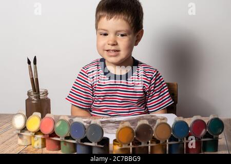 Un petit garçon de 2-3 ans dans un t-shirt rayé multicolore dessine des peintures guasya sur papier. Loisirs et divertissements pour les enfants, développement de l'enfant Banque D'Images